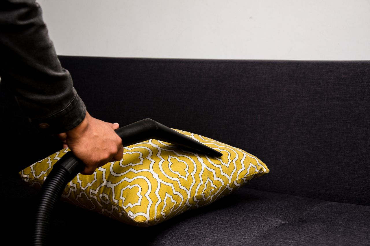 A close-up view of someone vacuuming a patterned pillow on a dark sofa indoors.
